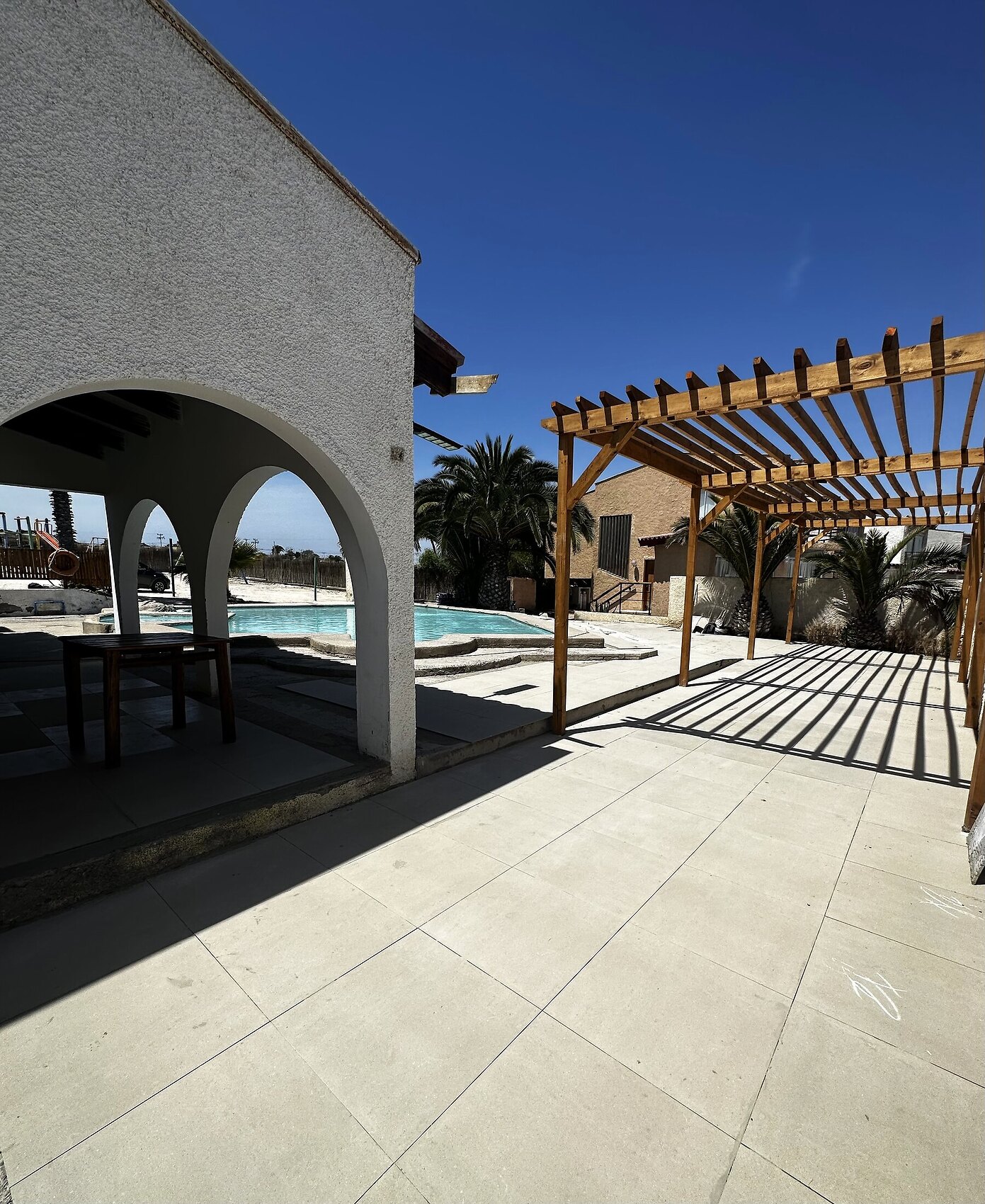Arquería blanca, pérgola de madera y patio pavimentado junto a la piscina del recinto